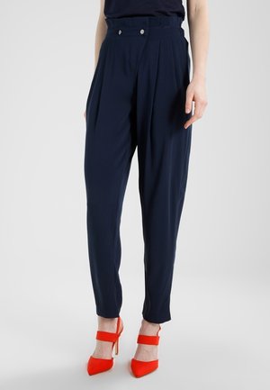 Femme portant un pantalon plissé taille haute bleu marine et des chaussures à talons hauts rouge vif à bout pointu avec des brides à la cheville.