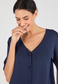 Top de color azul marino de manga corta con un profundo escote en V y cierre de botones. Tejido suave y texturizado con un ajuste relajado. Detalles de diseño minimalistas.