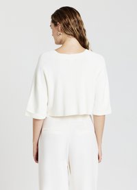 Maglione bianco corto con maniche ampie e arrotolate e scollatura ampia, in tessuto a maglia testurizzato. Indossato sopra pantaloni chiari.