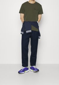 Olivgrön t-shirt med bröstficka, marinblå sweatpants och lila sneakers med vita och gröna accenter, bärs avslappnat.