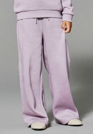 Persona che indossa pantaloni della tuta larghi color lavanda e un sudore coordinato, in piedi su un pavimento grigio, con scarpe beige visibili.