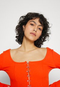 Top laranja com nervuras, decote quadrado e detalhes em metal em forma de laço. Possui mangas longas e um design justo.