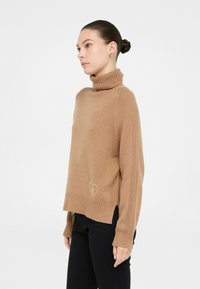 Donna con capelli scuri raccolti in uno chignon che indossa un dolcevita cammello con maniche a coste e pantaloni neri, in posa di profilo laterale.