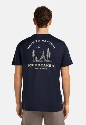 Hombre con camiseta azul marino que muestra una escena de camping en blanco, árboles, luna y el texto "Move to Natural ICEBREAKER Since 1995" en la parte trasera.
