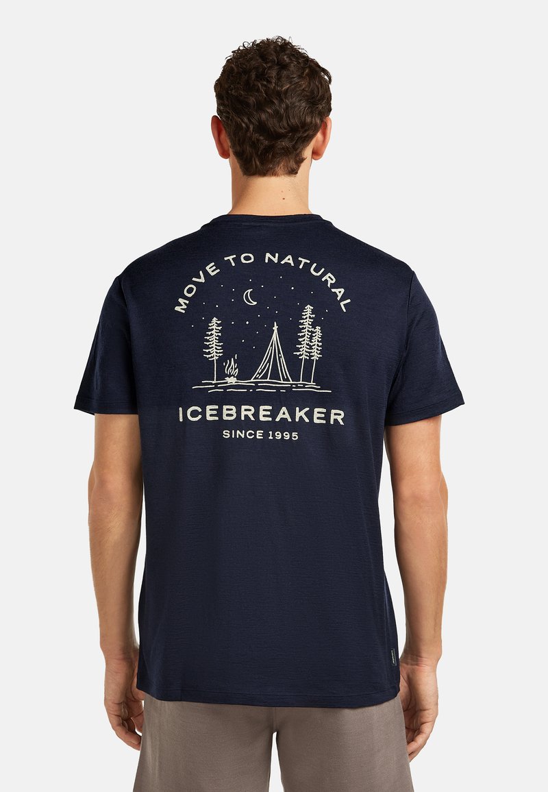 Hombre con camiseta azul marino que muestra una escena de camping en blanco, árboles, luna y el texto "Move to Natural ICEBREAKER Since 1995" en la parte trasera.