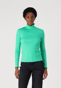 Green long-sleeve turtleneck top with a ruffled collar and cuffs; made of a smooth, stretchy fabric. Paired with dark jeans.