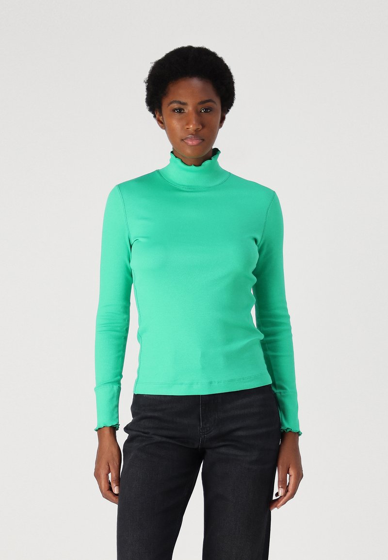 Green long-sleeve turtleneck top with a ruffled collar and cuffs; made of a smooth, stretchy fabric. Paired with dark jeans.