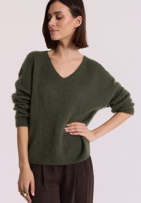 Oliwkowa, oversize sweter w serek wykonany z miękkiego, puszystego materiału z wywiniętymi rękawami; w zestawieniu z ciemnobrązowymi spodniami.