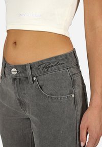 Zbliżenie na osobę w szarych dżinsach i skróconej białej bluzce z prążkami, odsłaniającej brzuch oraz z widocznymi detalami marki na odzieży.