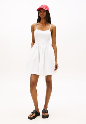 Γυναίκα που φοράει λευκό αμάνικο φόρεμα μήκους μέχρι το γόνατο, μαύρα σανδάλια πλατφόρμα και κόκκινο καπέλο, στέκεται μπροστά σε ένα απλό λευκό φόντο.