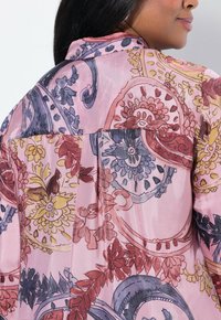 Leichtes, transparentes Blusenoberteil in Pink mit aufwendigen Blumen- und Paisleymustern in Rot-, Blau- und Gelbtönen; Kragendetails sind sichtbar.