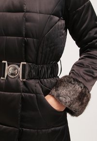 Černý vyp paddingovaný kabát s prošívaným designem, elastickým pasem s kovovou stříbrnou sponou a umělou kožešinou na manžetách.