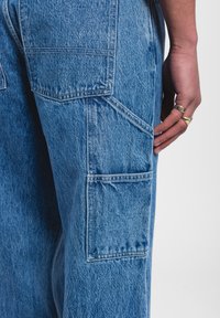 Pantalones cargo de mezclilla azul con múltiples bolsillos, textura pesada y acabado desgastado. Los detalles de costura visibles realzan el diseño.