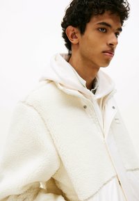 Weiße Fleecejacke mit plüschiger Textur, mit Reißverschluss und Druckknöpfen, über einem weißen Hoodie und einem grün gemusterten Hemd getragen.