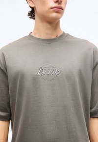 Νεαρό άτομο που φοράει μια απαλού πράσινου αποχρώματος μπλούζα με ένα διακριτικό κεντητό λογότυπο "Los Angeles Lakers" στο στήθος, απλό φόντο.