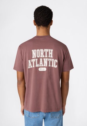 Mann trägt ein verblichenes lila T-Shirt mit dem Schriftzug „NORTH ATLANTIC“ und kleinem Champion-Logo auf dem Rücken, kombiniert mit hellblauen Jeans.