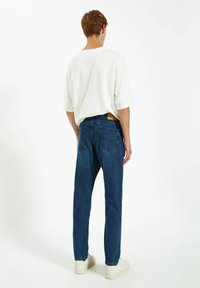 Camiseta blanca de algodón holgada con cuello redondo, combinada con jeans de mezclilla azul oscuro que cuentan con bolsillos traseros visibles y un parche de cuero.