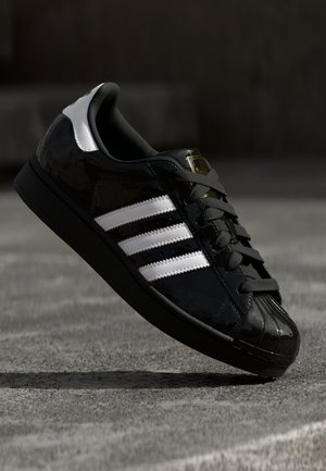 Sort sneaker med hvide striber og snørebånd, svævende over en tekstureret grå overflade i svagt lys.