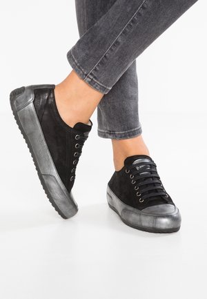 Czarne i srebrne sneakersy na sznurowadła noszone z podwiniętymi ciemnoszarymi dżinsami na skrzyżowanych nogach na białym tle.