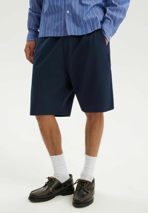 Mężczyzna w granatowych szortach, niebieskiej koszuli w paski, białych skarpetach do kostek i czarnych skórzanych sznurowanych butach, stojący z rękami w kieszeniach.