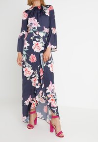 Kwiatowa sukienka maxi w granatowym kolorze z różowymi i białymi kwiatami. Długie rękawy, falbaniasty dół, podkreślona talia, w połączeniu z różowymi sandałami na słupku.