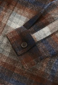 Chemise en flanelle à motif écossais dans des tons de marron, bleu marine et gris. Équipée d'un poignet à boutons et d'un tissu doux et texturé.