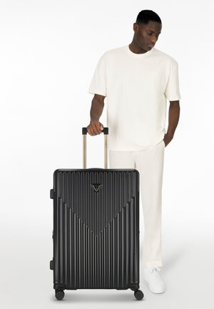 Valise rigide noire avec texture à arêtes verticales, quincaillerie en argent, poignée rétractable et quatre roues pour la mobilité.