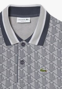 Polo Lacoste classica in grigio, con motivo geometrico, colletto a righe blu navy, due bottoni e logo ricamato verde.