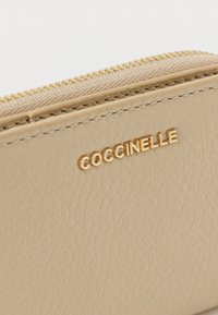 Beigenvärinen nahkalompakko, jossa on teksturoitu pinta ja kullanväriset metalliosat. Brändin nimi "Coccinelle" on painettu kohopainatuksena kullanvärisenä.