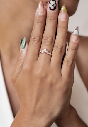 Rose gold ring with a wavy design, featuring small clear gemstones resembling leaves, worn on a hand with colorful, nail art.