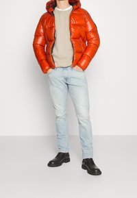 Puffjacke aus glänzendem orangefarbenem Material mit schwarzem Innenfutter, kombiniert mit hellblauen Jeans und schwarzen Stiefeln. Das Model steht mit den Händen in den Taschen.