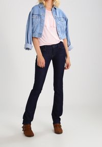 Jasnoniebieska kurtka dżinsowa z dwoma kieszeniami na piersi. Różowa bluzka z falbanami, ciemnoniebieskie dżinsy i brązowe botki za kostkę z frędzlami.