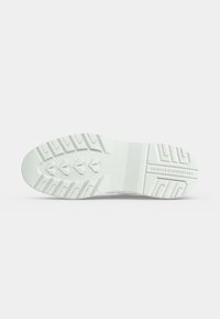 Semelle de chaussure blanche avec des rainures profondes et des motifs triangulaires, affichant le texte "COLE HAAN STUDIO" dans la zone du talon.