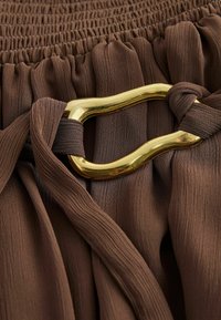 Tissu marron plissé rassemblé par une ceinture élastique avec une boucle en métal doré texturée sécurisant une ceinture assortie en tissu.