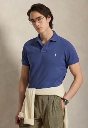 Jeune homme avec des lunettes portant un polo bleu, un pantalon à carreaux, une montre-bracelet et un pull crème noué autour de la taille, debout devant un fond uni.