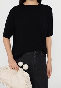 Haut en maille noir à manches courtes porté avec un jean foncé. Pochette en tissu blanc texturé avec une fermeture circulaire unique tenue dans une main.