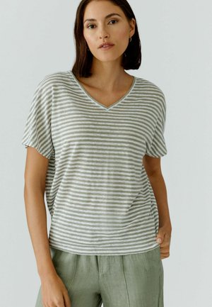 Woman with short dark hair wearing a grey and white striped V-neck T-shirt and light green pants, standing against a plain background.