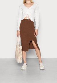 Bílý žebrovaný top s dlouhým rukávem a nabráním v přední části, spárovaný s hnědou žebrovanou pletenou sukní s bočním rozparkem. Béžová pruhovaná taška a tenisky.