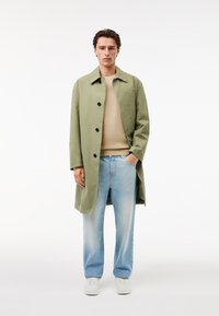 Mann in hellgrünem Mantel über beigem Pullover, hellblauen Jeans und weißen Sneakers, stehend vor einem schlichten weißen Hintergrund.
