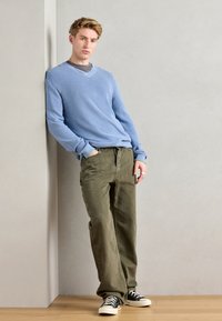 Jasnoniebieski sweter z dekoltem w serek, oliwkowe spodnie o luźnym kroju i czarne sneakersy. Zauważalny pierścionek na prawej ręce.