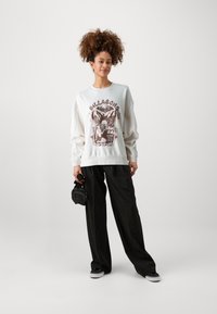 Felpa bianca con stampa grafica che presenta testo e immagini. Abbinata a pantaloni neri a righe e sneaker nere. Borsa piccola in mano.