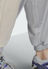 Gaiši pelēki un krēmkrāsas sporta bikses ar elastīgiem aplīšiem, apvienotas ar sudrabām un violetām sporta apaviem ar sietu augšdaļu un šnūrēm.