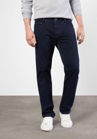 Jean en denim bleu foncé avec une coupe droite, des détails de couture visibles et une fermeture à bouton à la taille, associés à des baskets blanches.