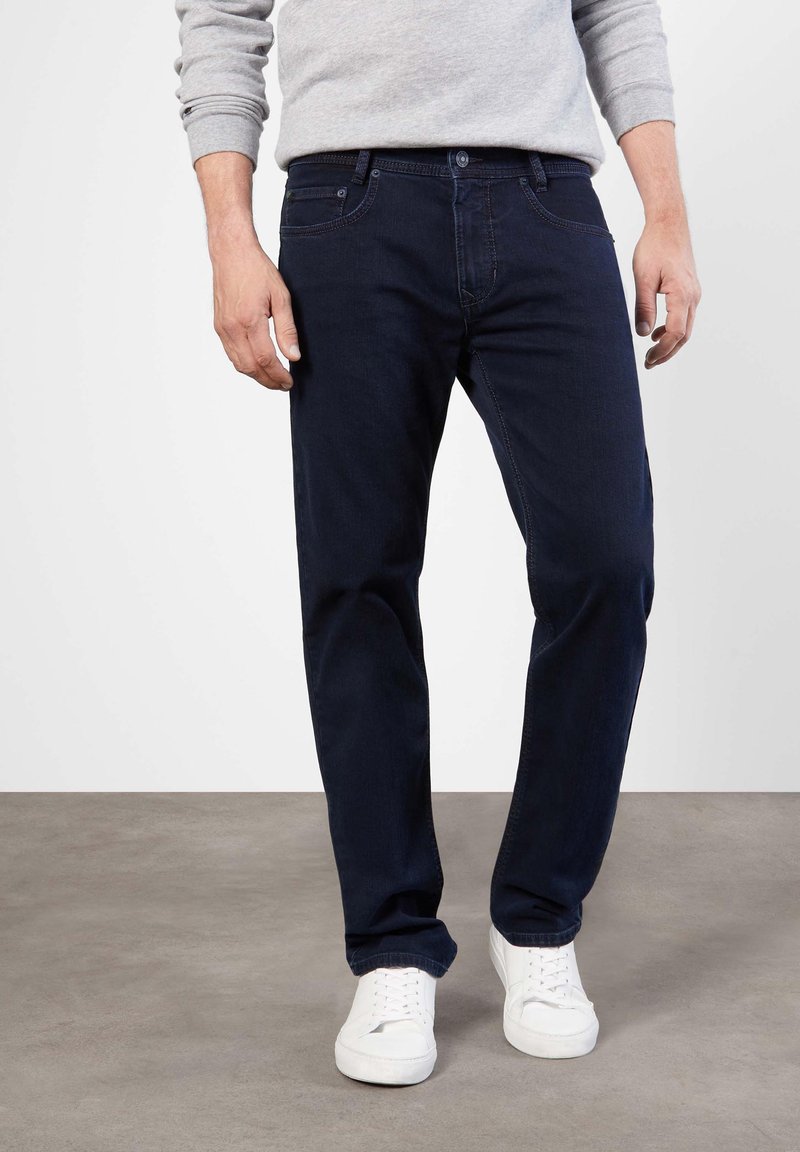 Jean en denim bleu foncé avec une coupe droite, des détails de couture visibles et une fermeture à bouton à la taille, associés à des baskets blanches.