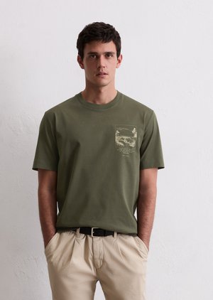 T-shirt en coton vert avec col rond, manches courtes, présentant un imprimé de poche avec des détails de paysage, associé à un pantalon beige.