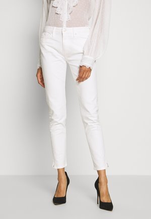 Femme portant un jean blanc taille haute associé à une blouse blanche transparente à manches longues et des chaussures à talons noirs, debout sur un fond uni.