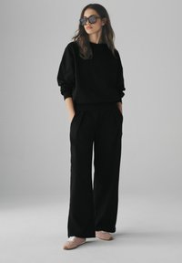 Felpa nera con scollatura rotonda e maniche lunghe, abbinata a pantaloni neri a gamba larga. Il modello indossa scarpe rosa e occhiali da sole oversize.