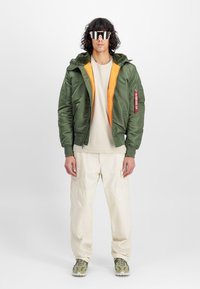 Green bomber jacket with orange lining, fuzzy collar, and pocket details; paired with cream cargo pants and green sneakers.