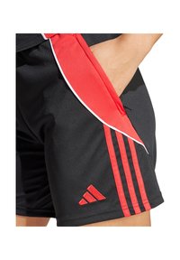 Mano poggiata nella tasca di pantaloni sportivi neri con strisce rosse e logo, con un dettaglio di rifinitura rosso e bianco.