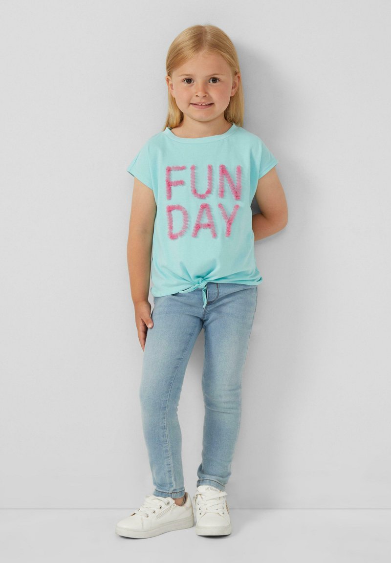 Hellblaues T-Shirt mit "FUN DAY" in flauschigen pinken Buchstaben, vorn geknotet, kombiniert mit hellblauen engen Jeans und weißen Sneakers.